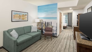A hotel room with a green sofa, a brown armchair, a flat-screen TV, and a large painting of a beach scene.