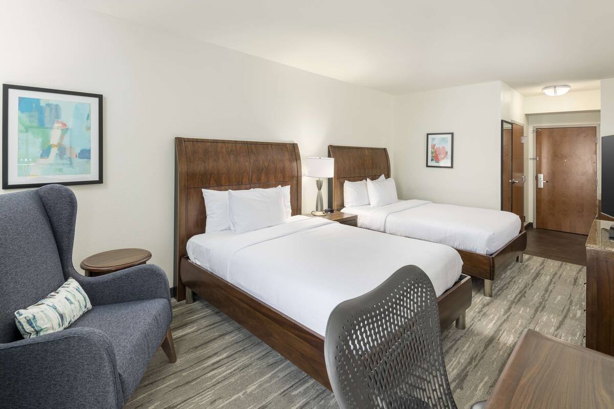 A hotel room with two beds, a grey armchair, a small table, and a TV.