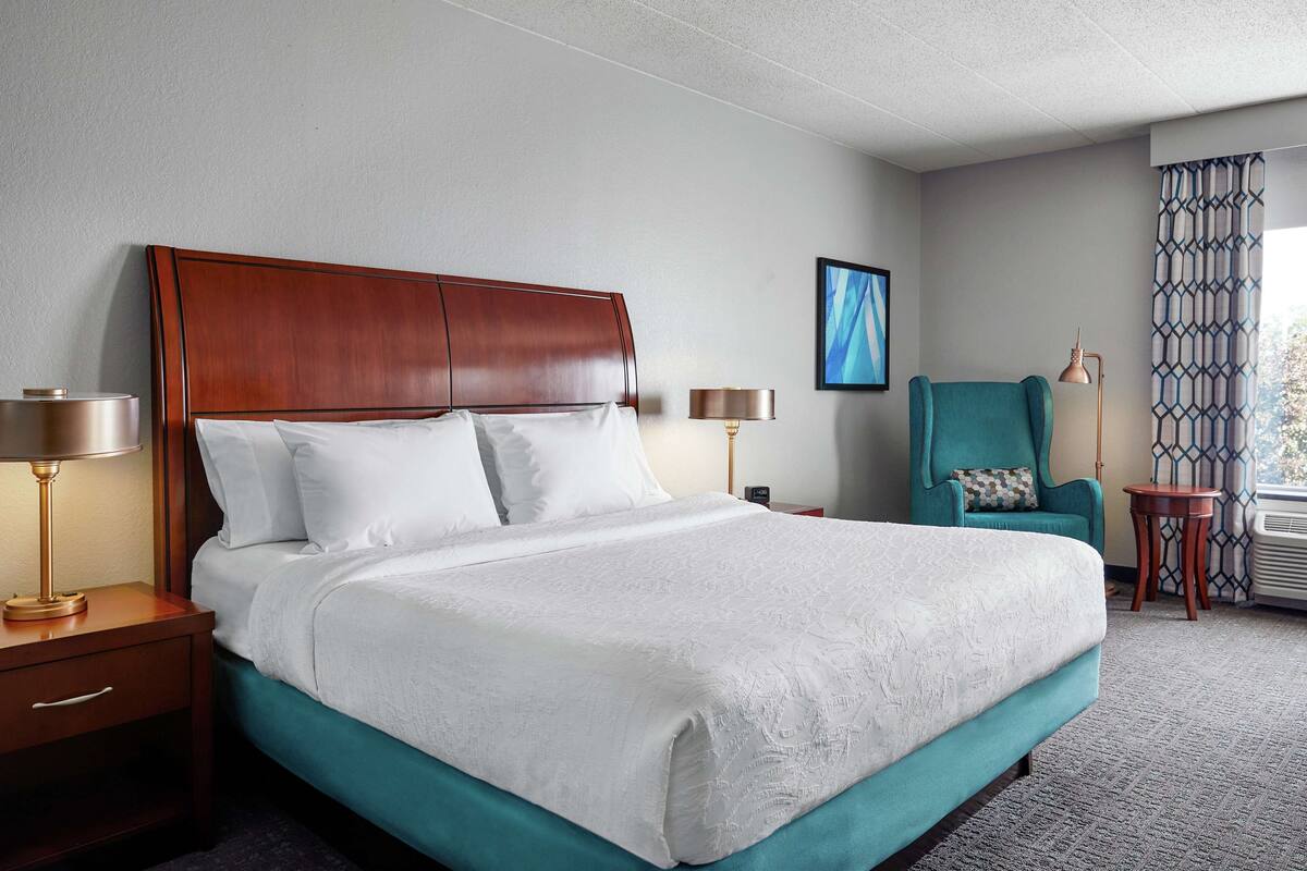Une chambre d’hôtel avec un grand lit, deux tables de chevet avec des lampes, un fauteuil turquoise et un rideau à motifs.