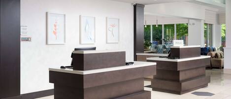 A modern hotel reception area with a clean, minimalist design, featuring a reception desk, framed artwork on the wall, and a view of the outdoor seating area.