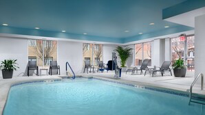 En indendørs swimmingpool med klart, blåt vand, omgivet af liggestole og potteplanter.