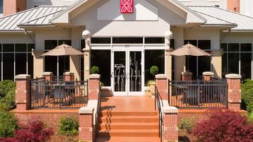 Entrada de hotel con logotipo rojo, puertas dobles y un corredor cubierto con sombrillas.