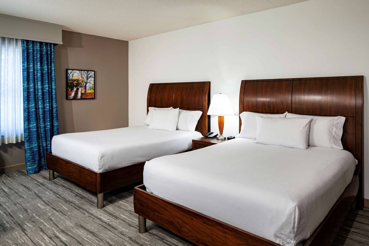 A hotel room with two beds, a nightstand with a lamp, a painting on the wall, and a window with blue curtains.