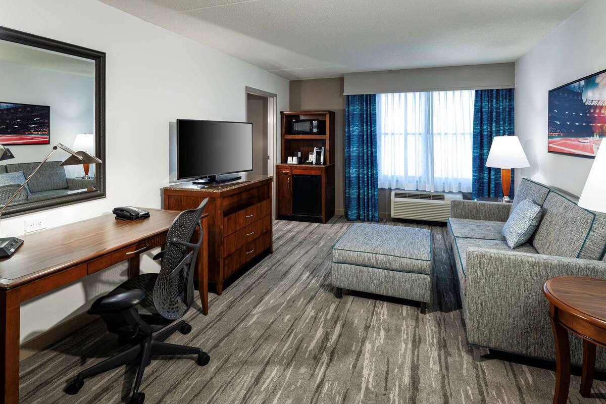 A hotel room with a desk, chair, television, sofa, and a small table with a lamp.