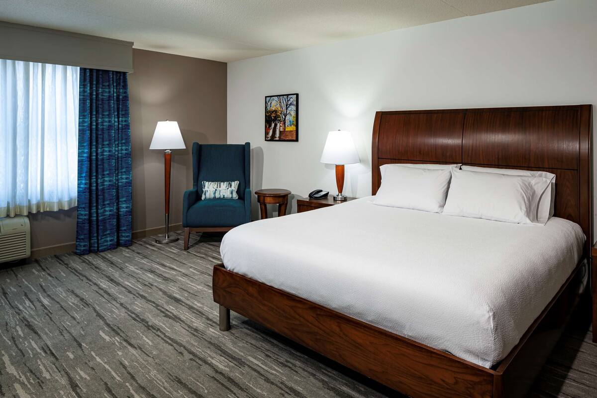 A hotel room with a large bed, two bedside lamps, a chair, a small table, and a painting on the wall.