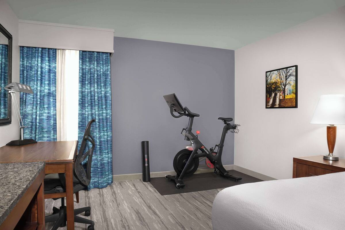 A hotel room with a bed, a desk with a chair, two exercise bikes, a lamp, and a painting on the wall.