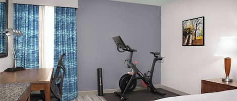A hotel room with a bed, a desk with a chair, two exercise bikes, a lamp, and a painting on the wall.