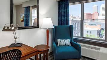 A hotel room with a large window, a teal armchair, a wooden desk, a lamp, and a mirror.