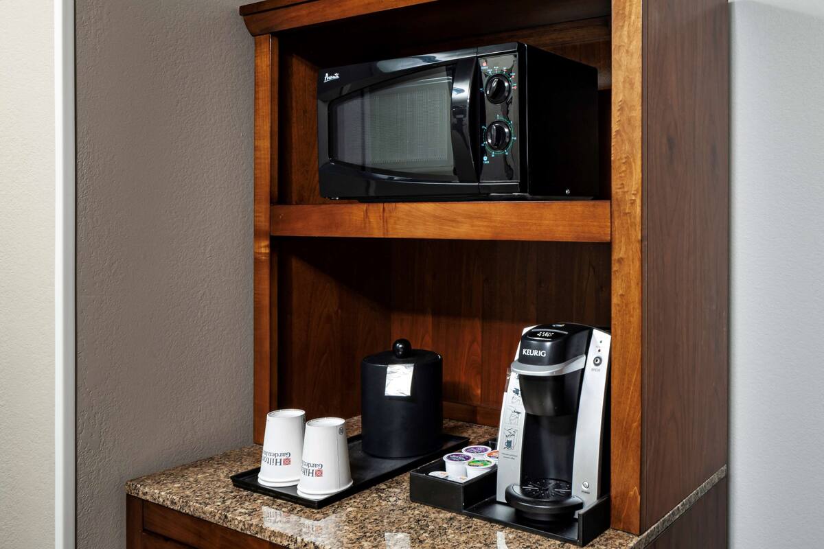 A kitchenette with a microwave, coffee maker, and two coffee cups on a granite countertop.