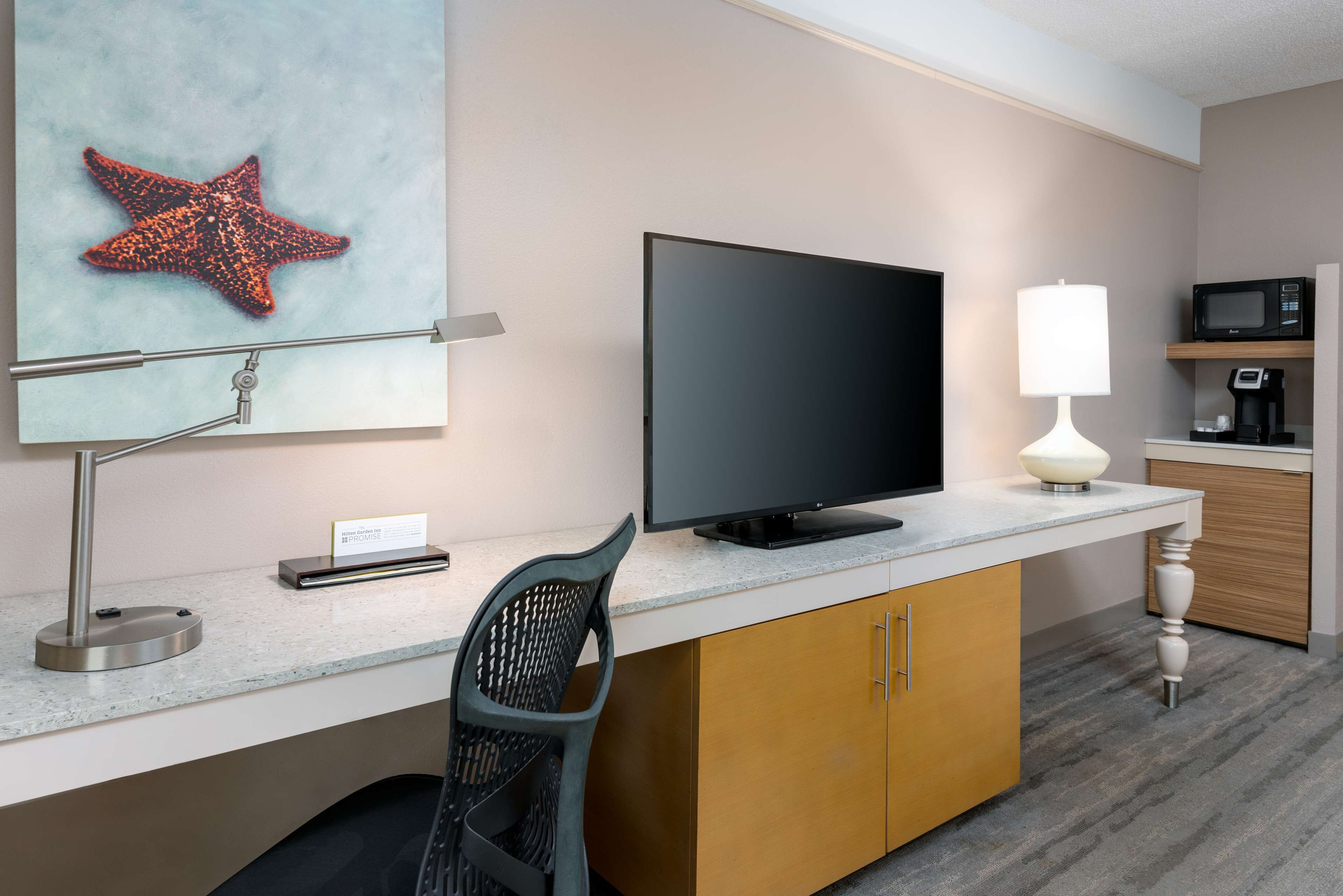 A modern hotel room with a flat-screen TV, a desk with a lamp, a chair, and a starfish decoration on the wall.