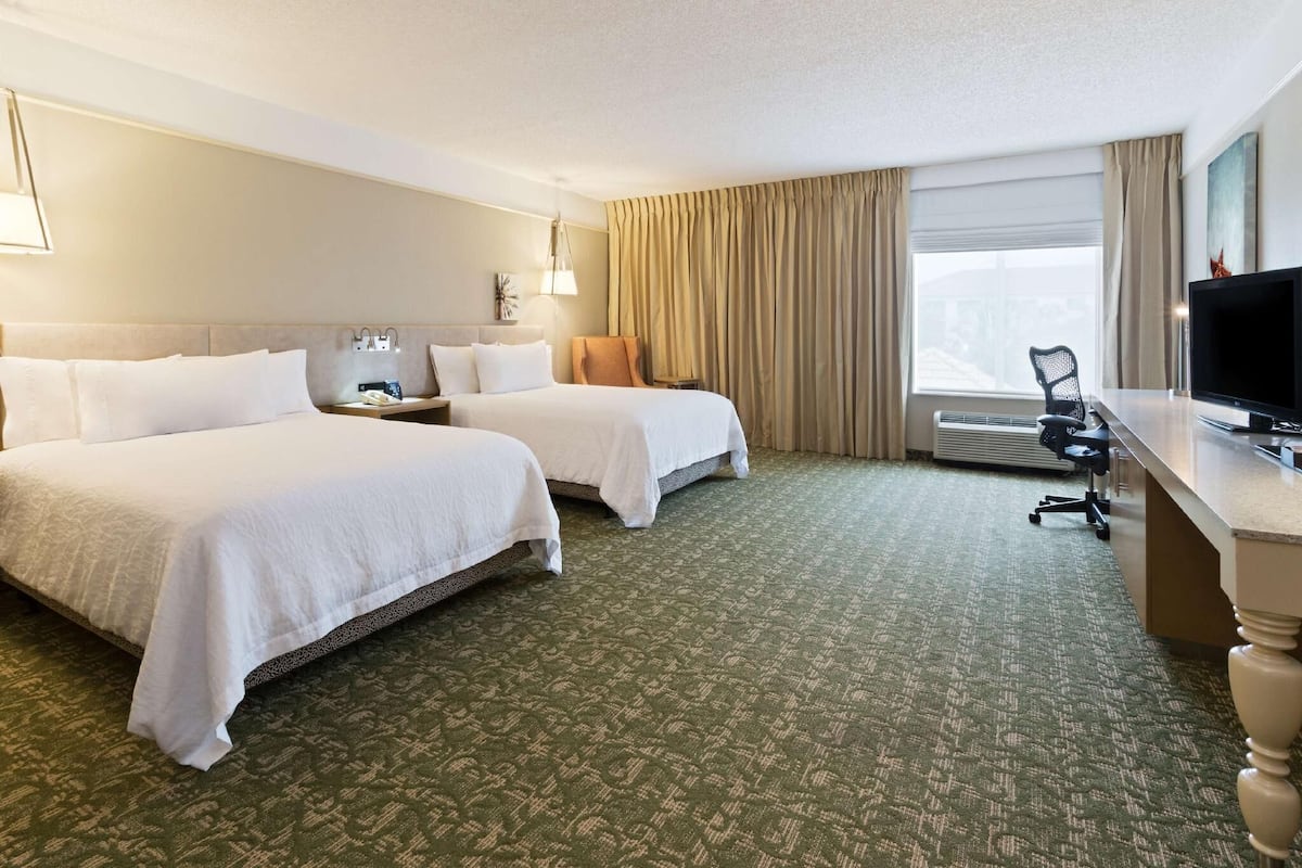A hotel room with two beds, a desk, a television, and a large window with curtains.