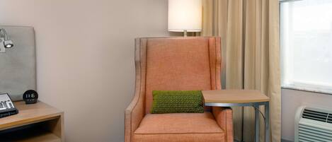 Habitación de hotel con un sillón individual, una mesita pequeña, una lámpara, una mesita de noche con teléfono y reloj, y una ventana con cortinas.