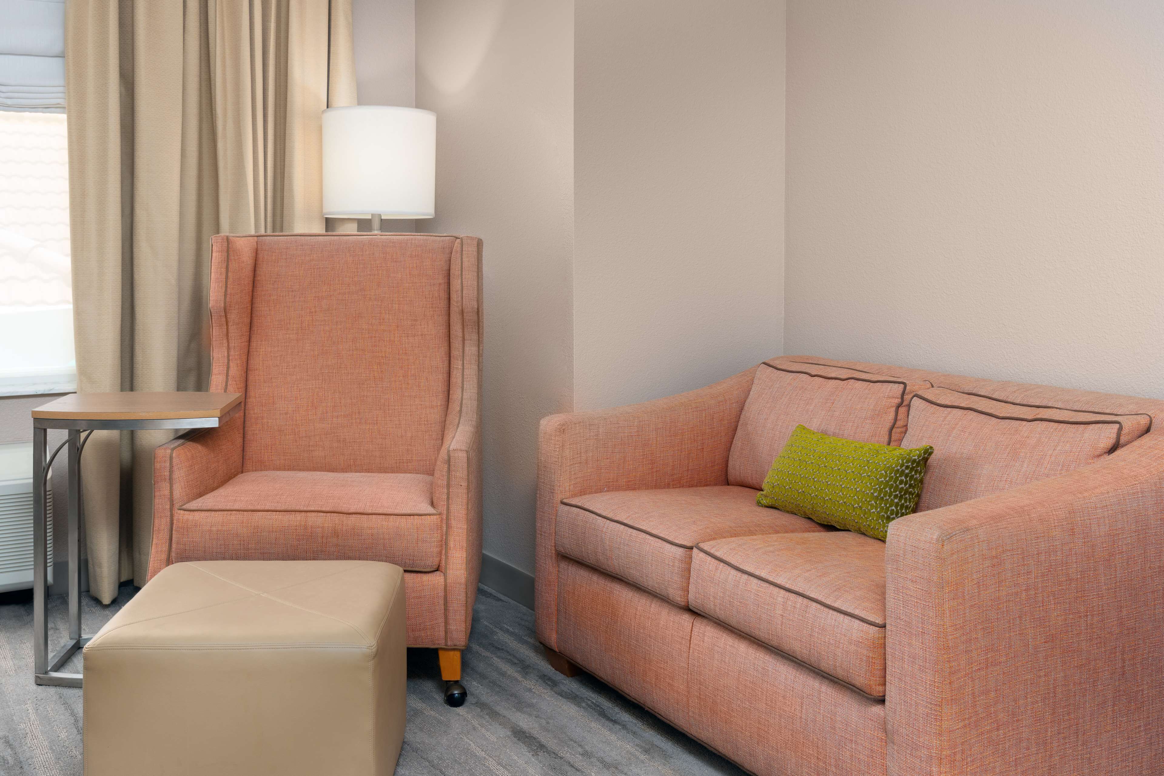 A hotel room with a beige ottoman, a pink sofa with a green pillow, a tall armchair, and a side table with a lamp.