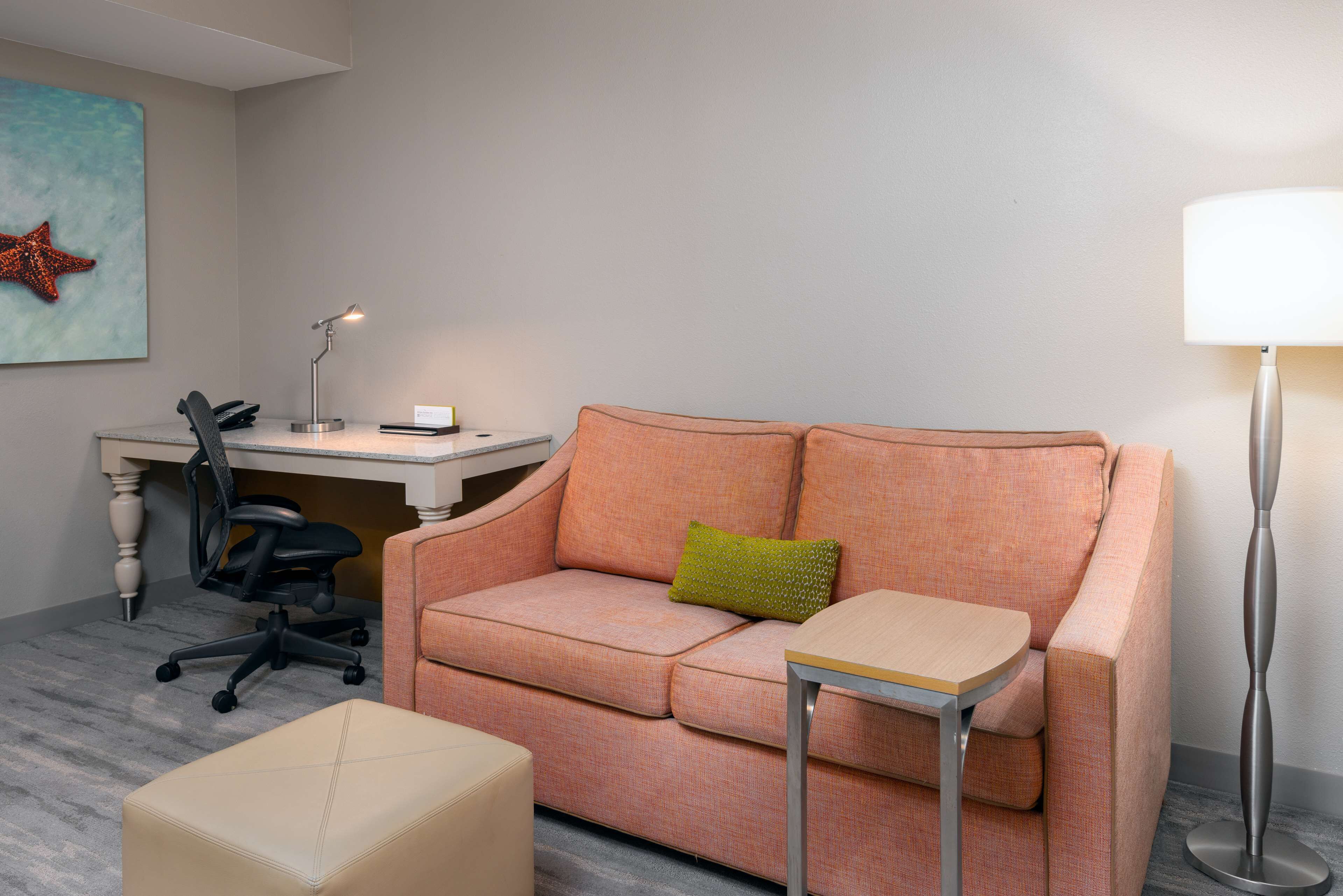 Et moderne hotelværelse med en lyserød sofa, et lille bord, et skrivebord med en computer, en stol og en lampe.