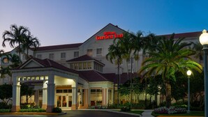 Um Hilton Garden Inn com arquitetura clássica, apresentando uma entrada destacada com área coberta e palmeiras em primeiro plano.