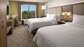 A hotel room with two beds, a desk, and a chair. There is a window with a view of trees and a lamp on the desk.
