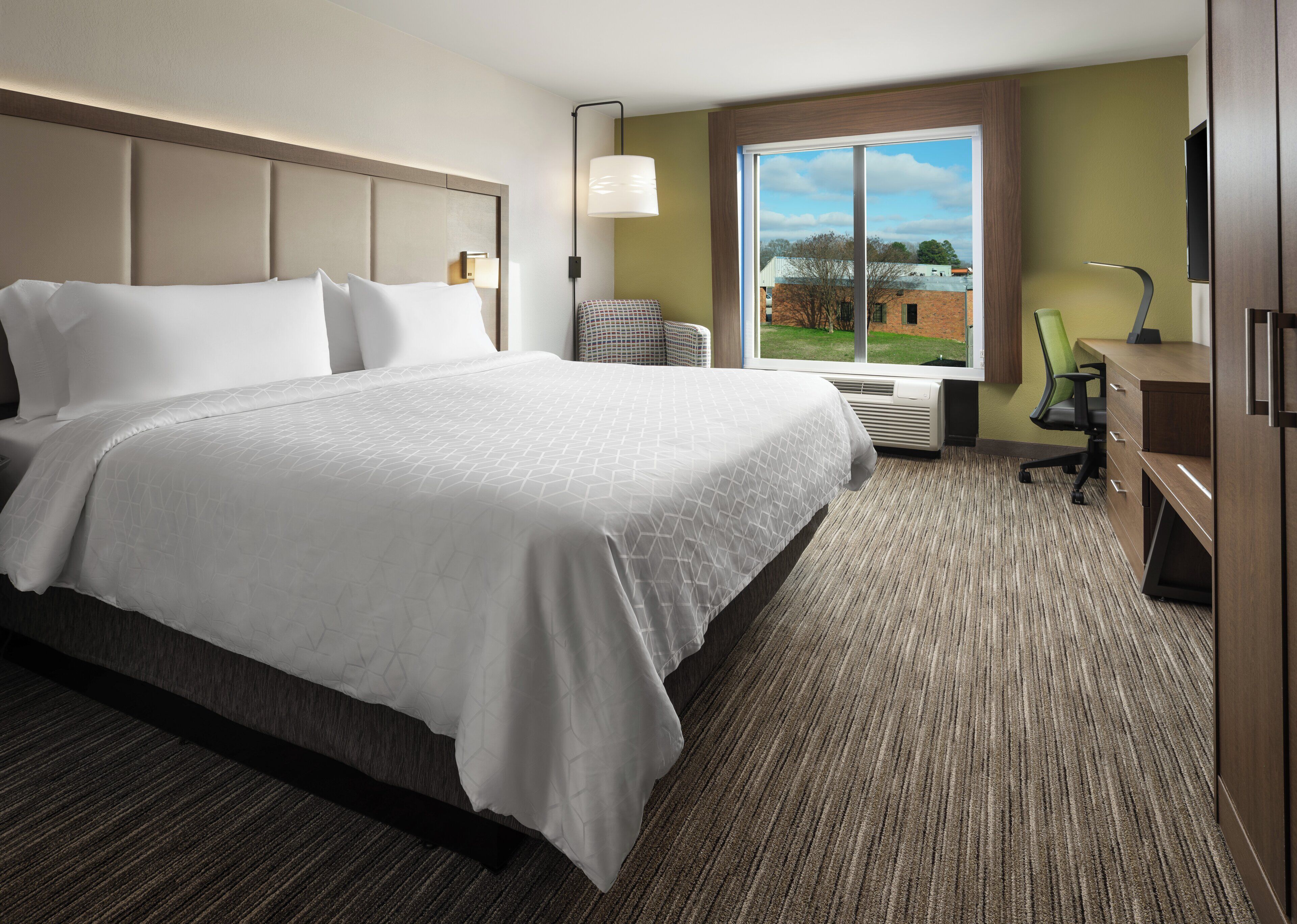 A hotel room with a large bed, a desk, a chair, a lamp, and a window with a view of buildings.
