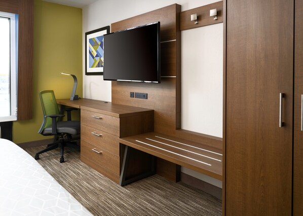 A hotel room with a wooden desk, a flat-screen TV, a green chair, and a bed with a patterned bedspread.