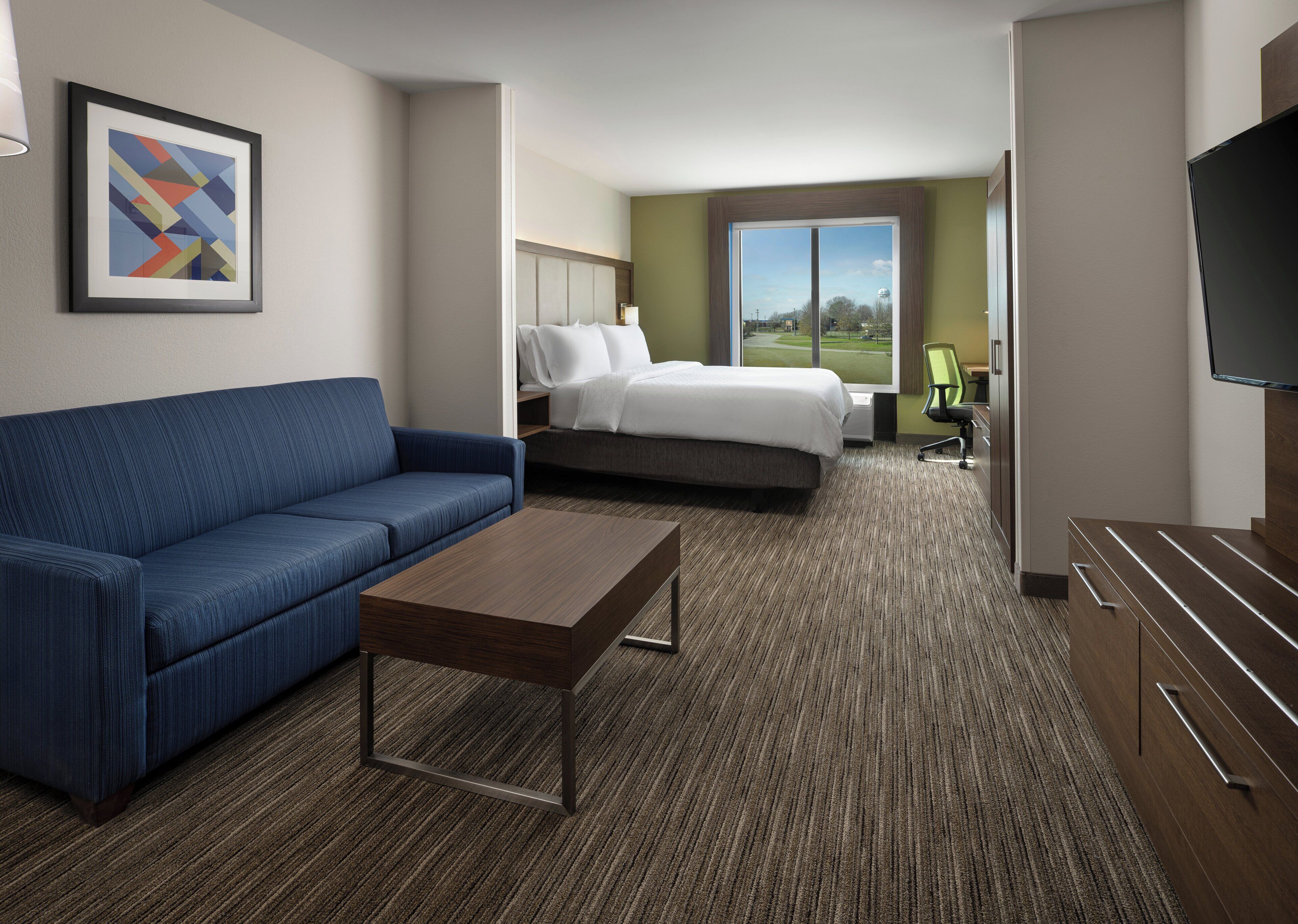 A hotel room with a blue sofa, a wooden coffee table, a bed with white linens, a green chair, and a TV on a wooden cabinet.