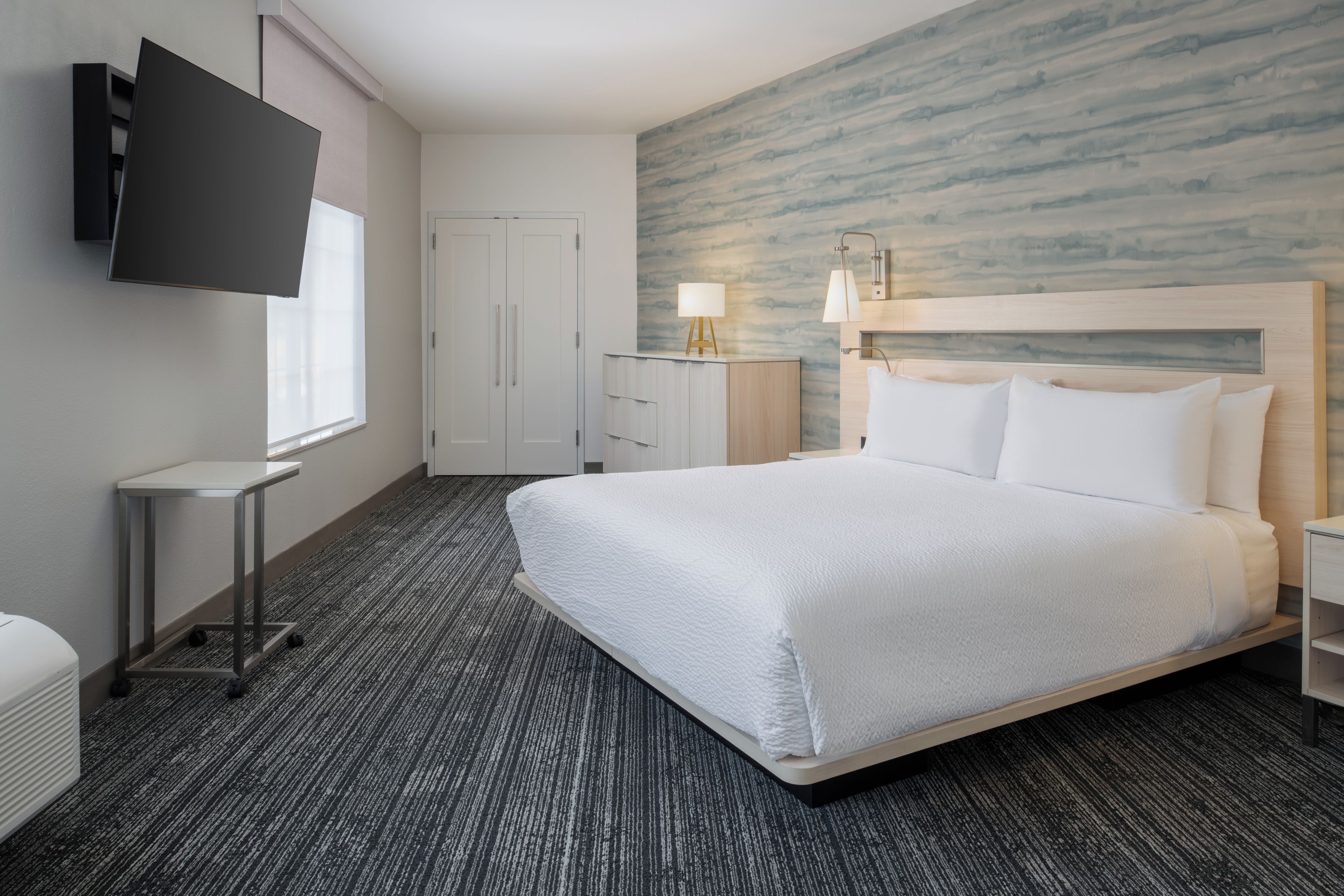 A hotel room with a large bed, a flat-screen TV mounted on the wall, a bedside table with a lamp, and a wardrobe.