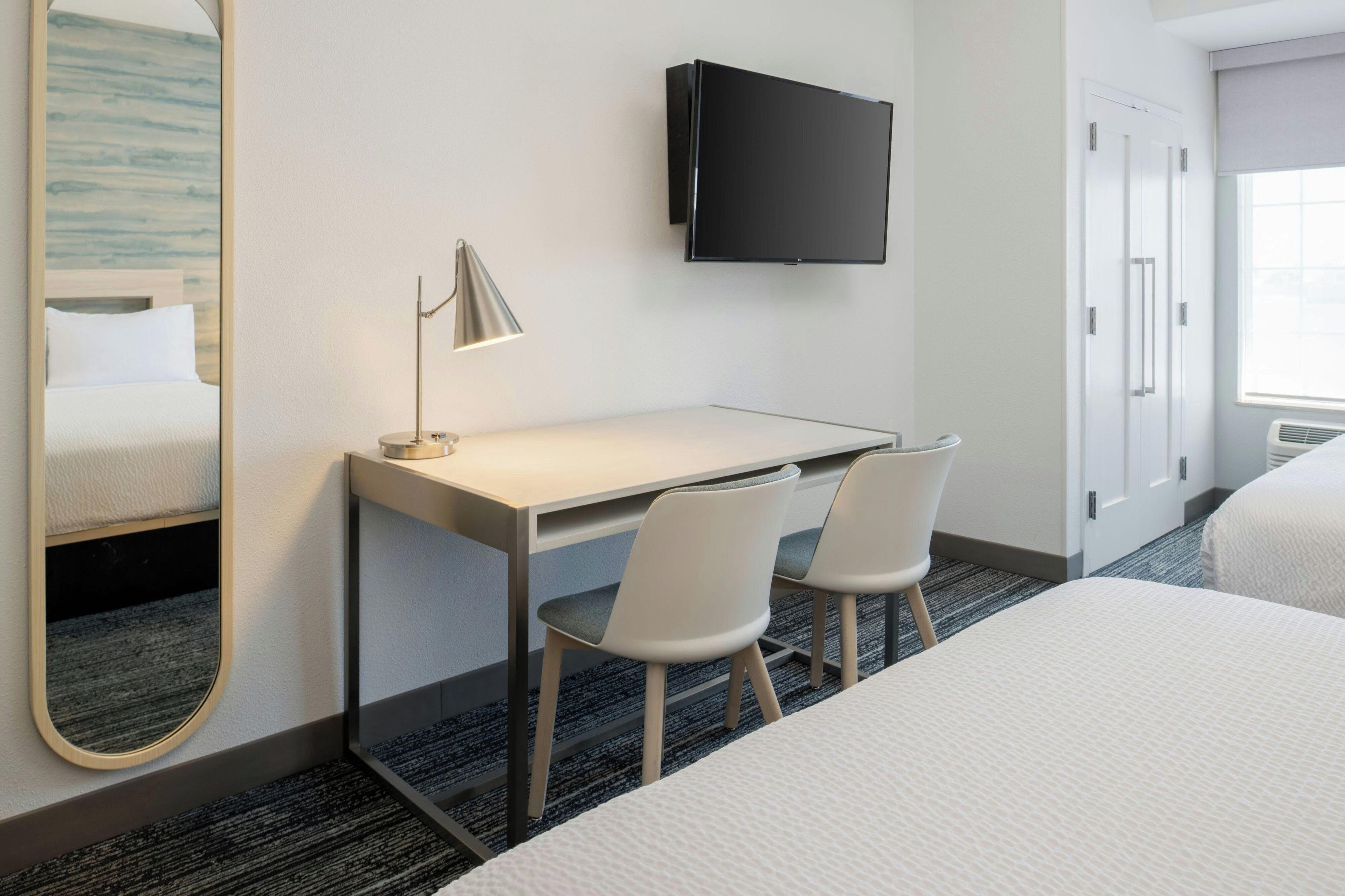 A hotel room with a bed, a desk with two chairs, a flat-screen TV, and a large mirror.