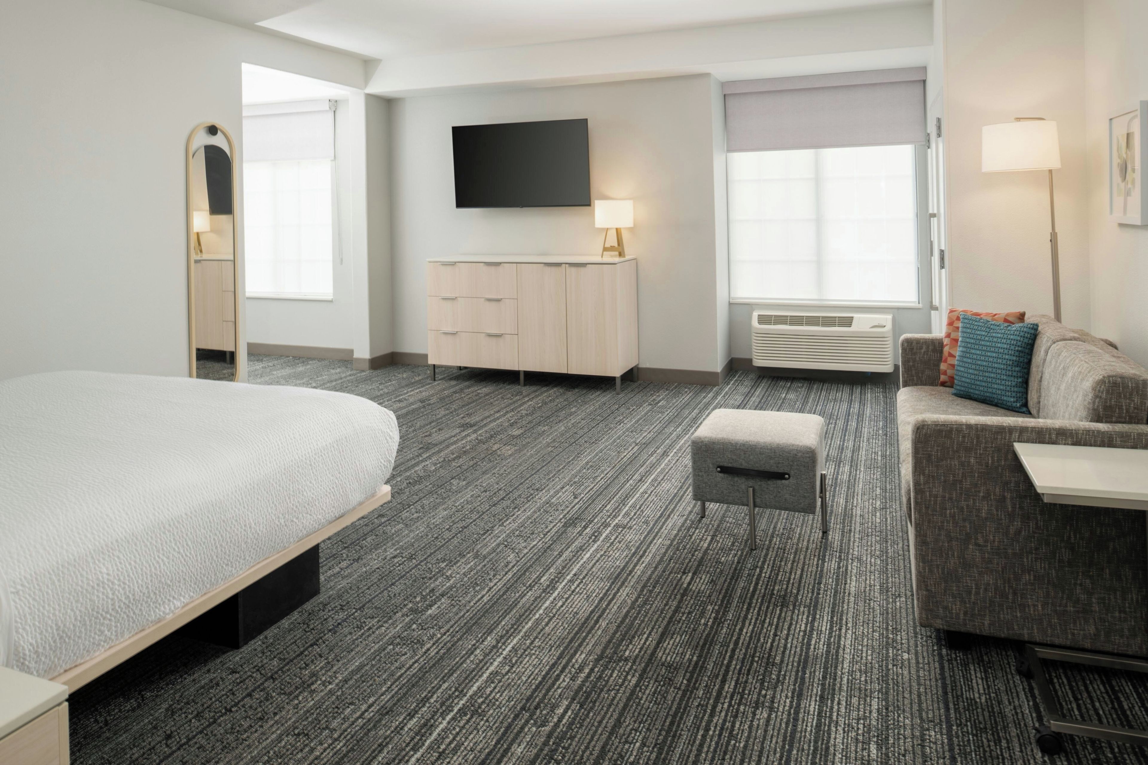 A hotel room with a bed, a sofa, a small table, a TV, and a window with blinds.