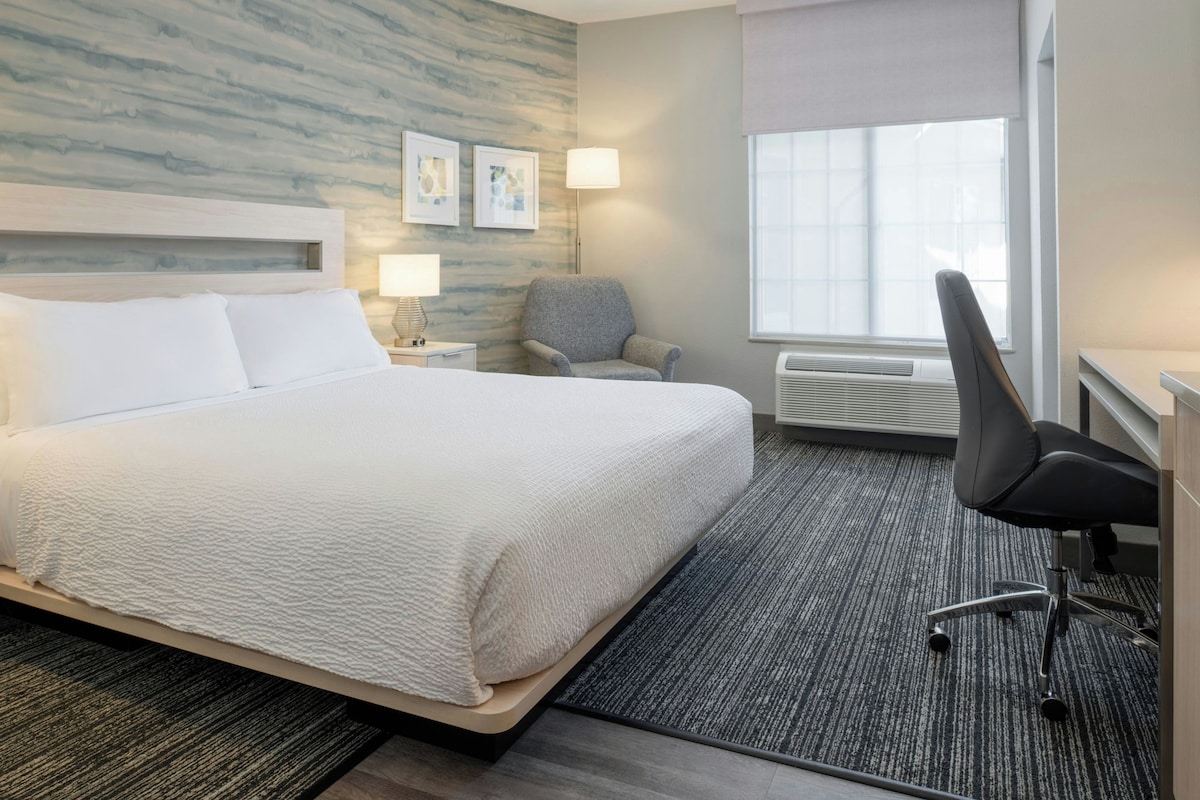 A hotel room with a large bed, a chair, a desk, a lamp, and a window.