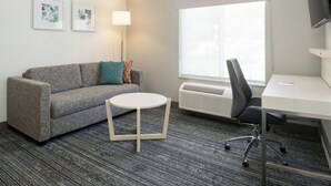 A hotel room with a grey sofa, a small round table, a desk with a chair, a standing heater, and a window with a view. - TownePlace Suites by Marriott San Pablo (San Pablo)