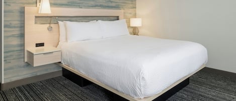 Quarto de hotel com cama, mesa de cabeceira, luminária na parede e piso coberto por carpete.