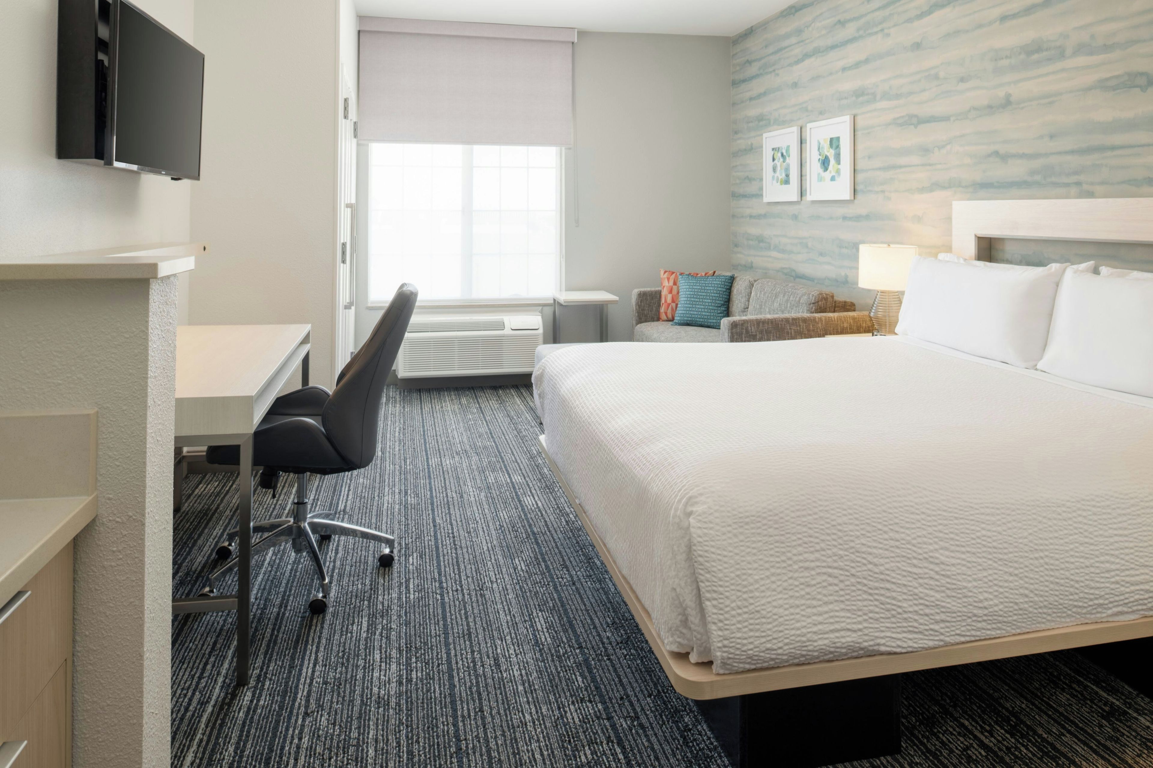A hotel room with a bed, a desk, a chair, a TV, a window with blinds, and artwork on the wall.