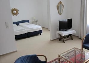 Hotellihuone, jossa on sänky, jalustalla oleva televisio, tuoli ja pöytä, jonka päällä on kuvioitu pöytäliina.