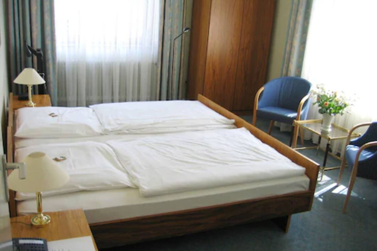 En dobbeltseng med hvide sengetøj, et natbord med lampe, en stol og et lille bord med en plante.
