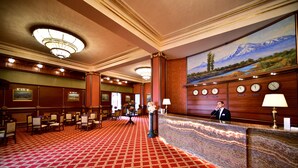 Un vestíbulo de hotel con alfombra roja, un mostrador de recepción, un cuadro de montañas y un reloj.