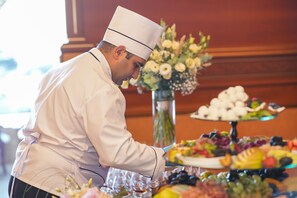 Um chef com uniforme e chapéu branco prepara comida num buffet com variedade de pratos e um arranjo floral.