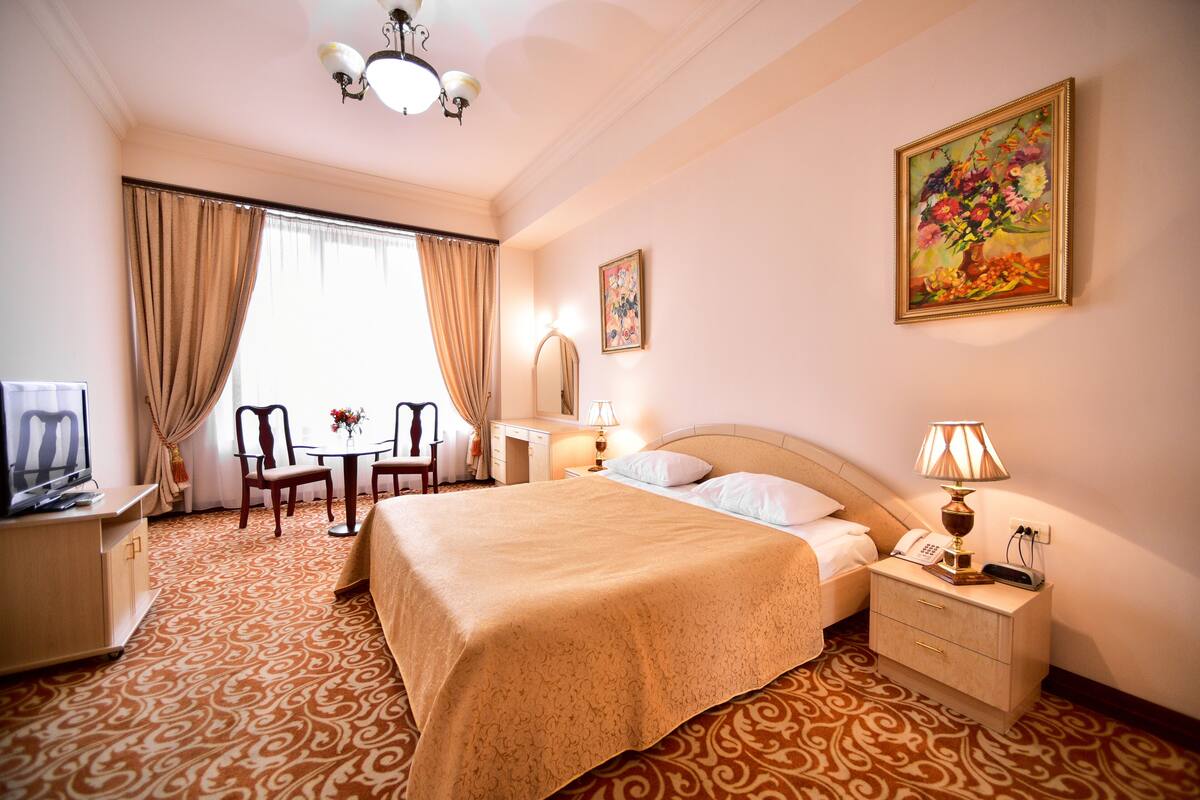 A hotel room with a large bed, a desk with a chair, a TV, a bedside table with a lamp, and a painting on the wall.