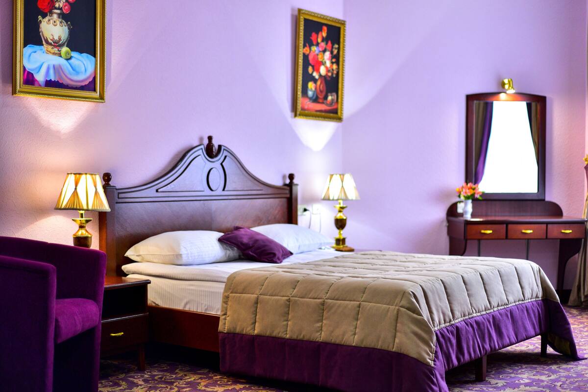 A hotel room with a wooden bed, purple and beige bedding, two bedside lamps, a purple armchair, a dresser with a mirror, and two wall-mounted paintings.