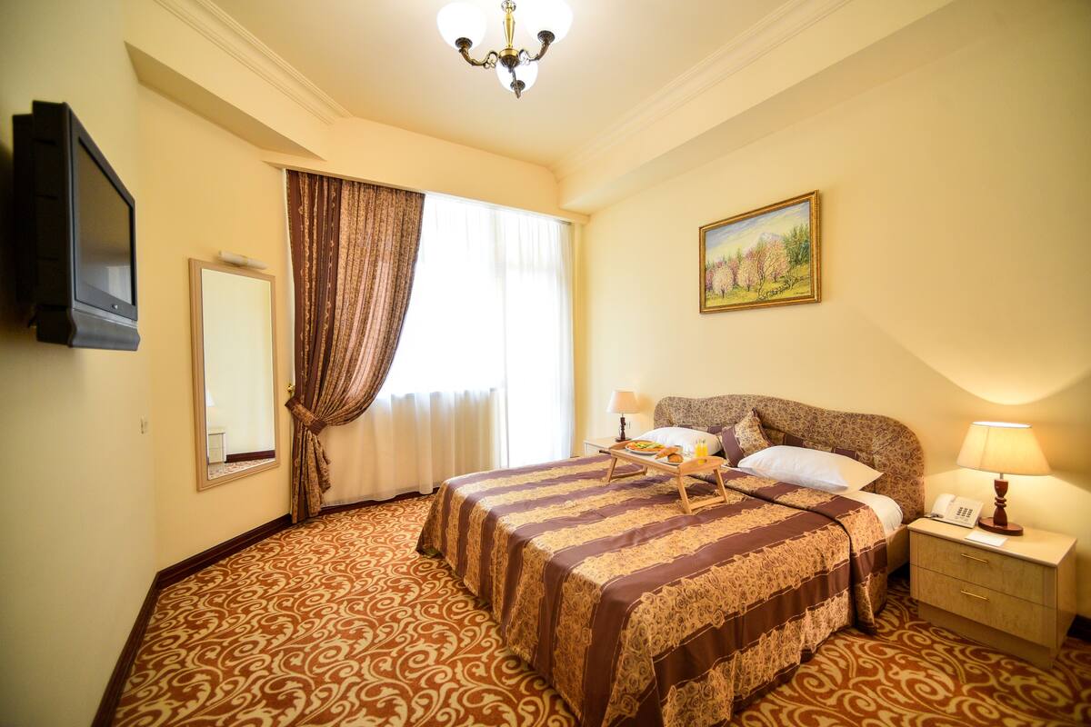 A hotel room with a large bed, a bedside table with a lamp, a painting on the wall, and a window with curtains.