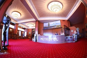 Un hall d’hôtel avec un tapis rouge, un comptoir d’accueil portant l’inscription « METROPOLIS », et un grand tableau représentant un paysage de montagne accroché au mur.