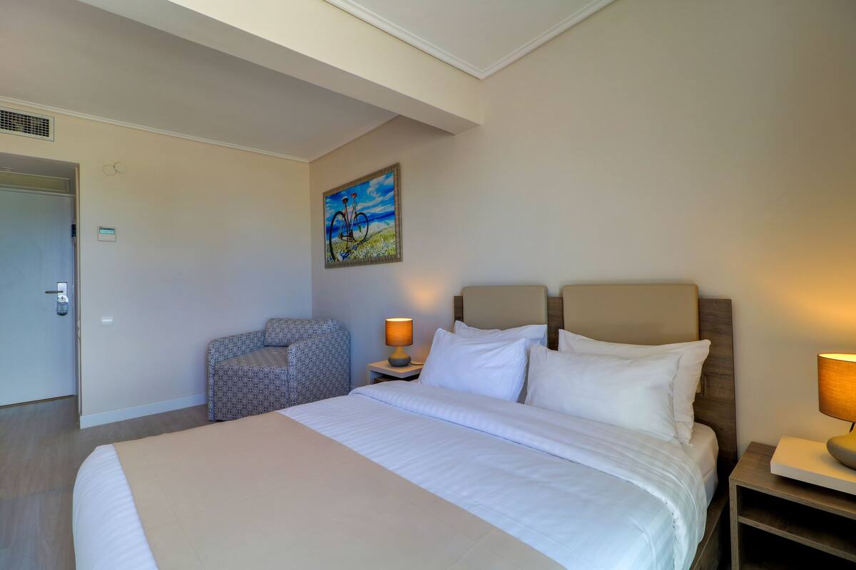 Ein Hotelzimmer mit einem großen Bett, einem Sessel, einem Nachttisch mit Lampe und einem Bild an der Wand.