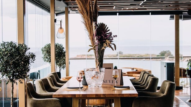 Een restaurant met een lange houten tafel, meerdere stoelen, een vaas met gedroogde planten, een fles en een glas wijn.