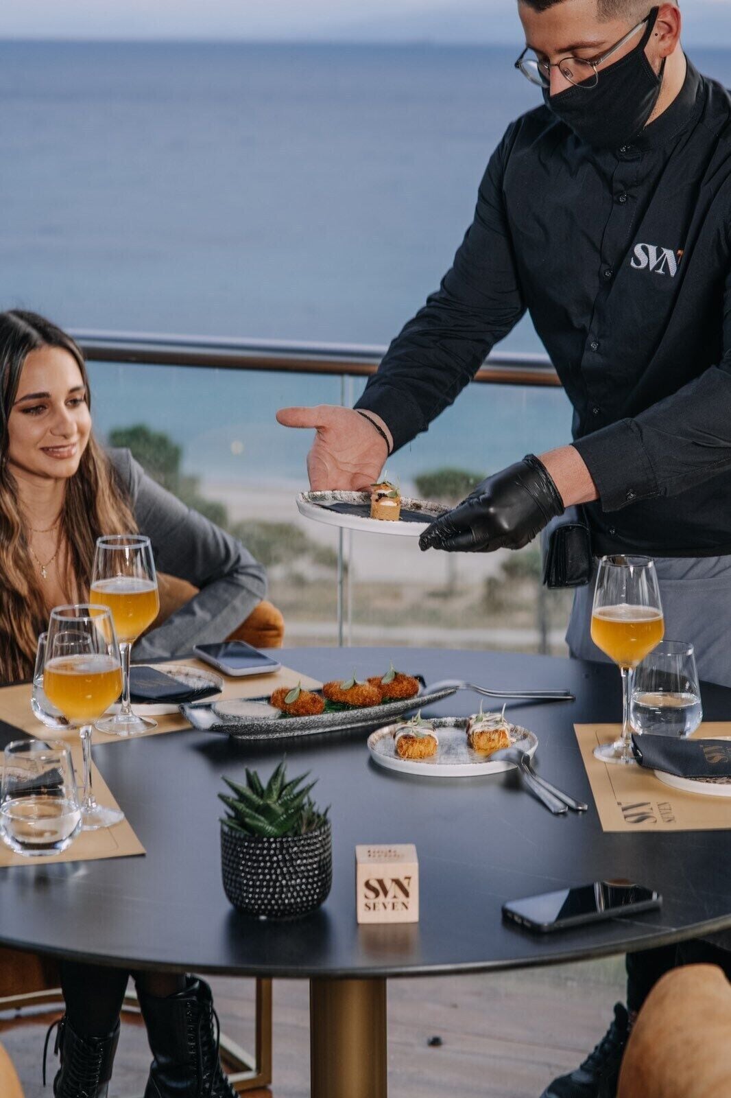 Una mesa de comedor con vista al mar, una persona sirviendo comida y un letrero que dice "SVN SEVEN".