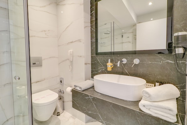 A modern bathroom with a glass-enclosed shower, a white toilet, a large oval sink, and a mirror above the sink. - Palace Hotel Athens Riviera (Glyfada)
