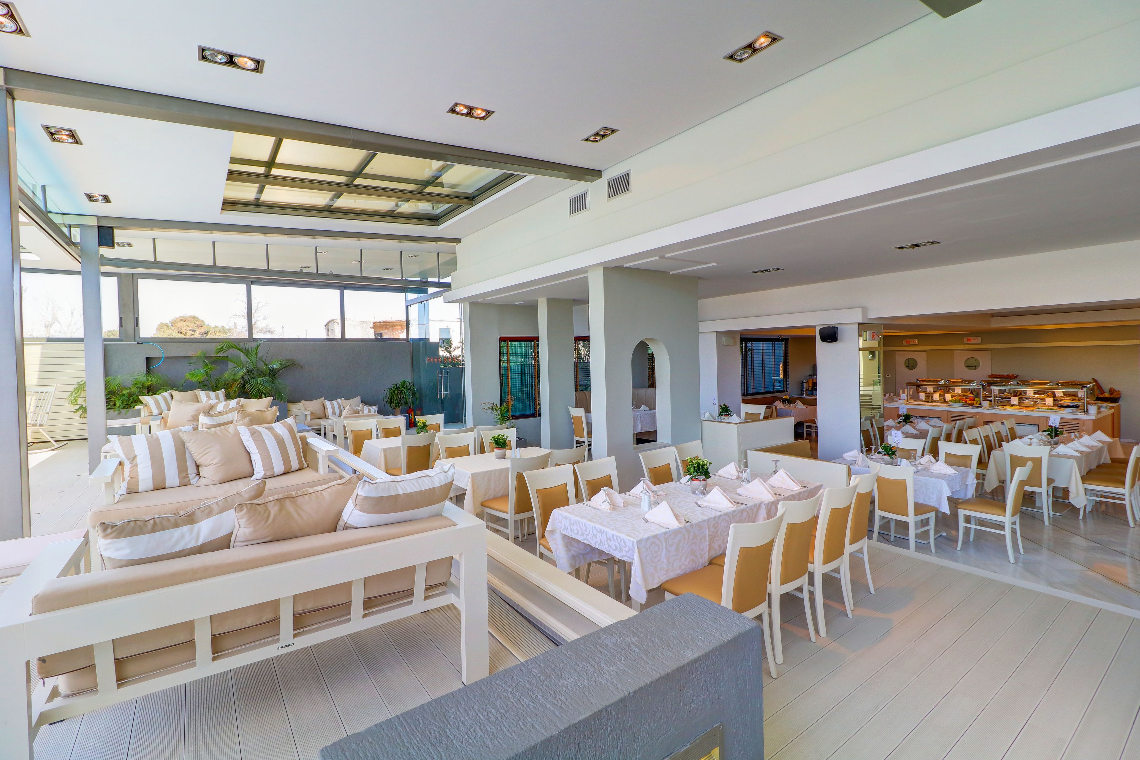 A spacious indoor dining area with multiple tables and chairs, a long white sofa, and a kitchen in the background.