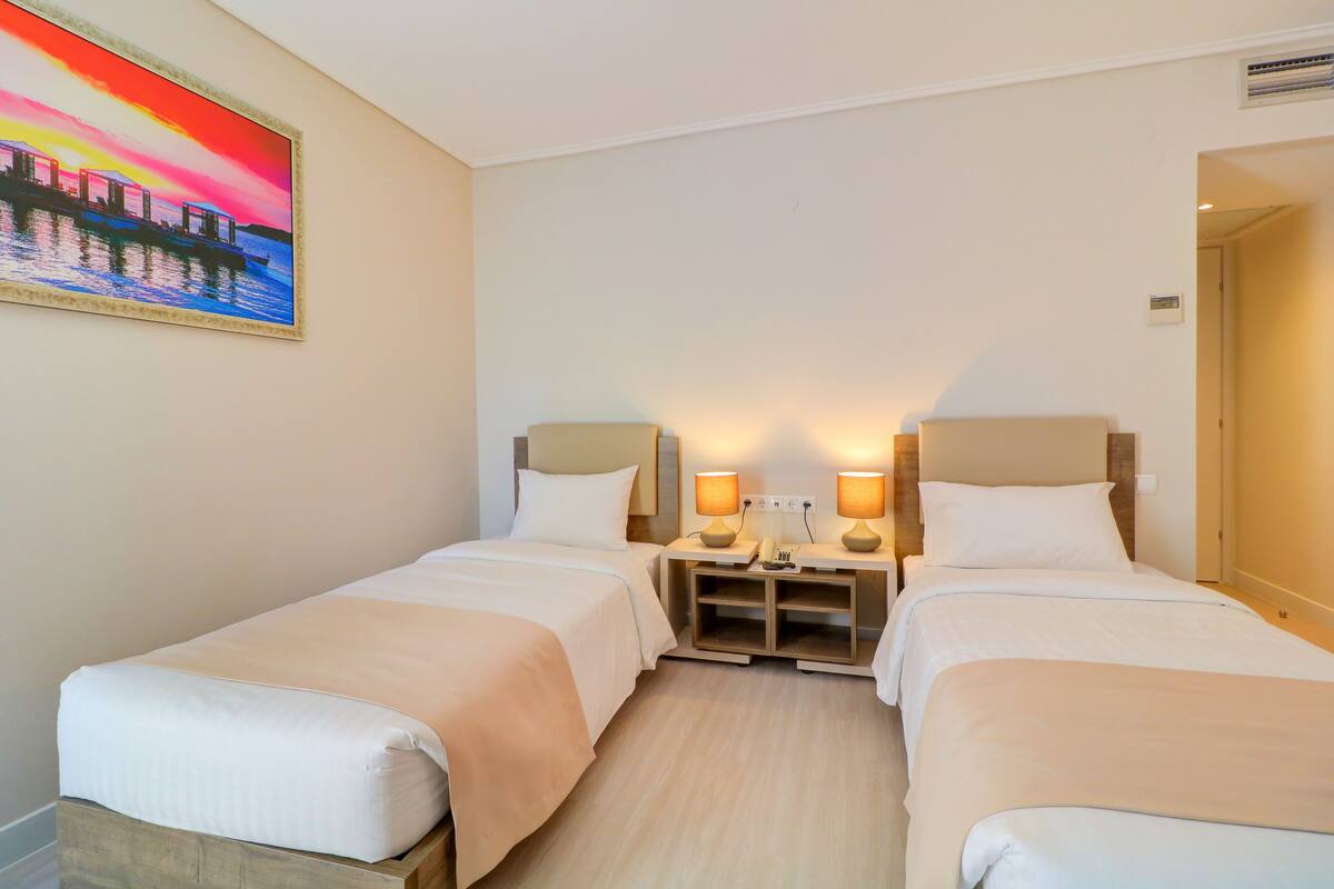 Ein Hotelzimmer mit zwei Betten, einem Nachttisch mit Lampen, einem Bild an der Wand und einer Tür rechts.