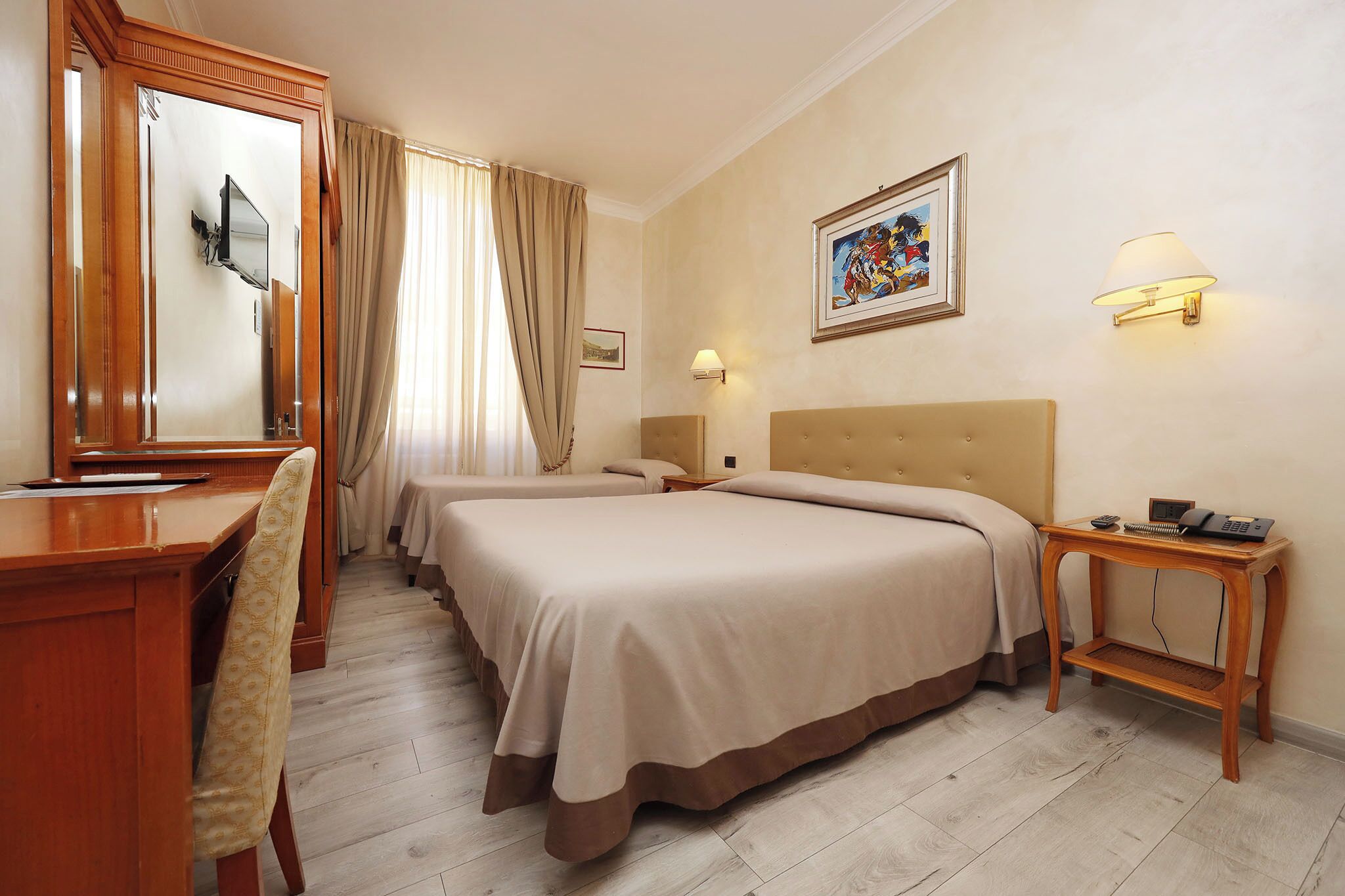Une chambre d’hôtel avec deux lits, un bureau, une chaise, un miroir, une table de chevet avec une lampe et un tableau encadré au mur.