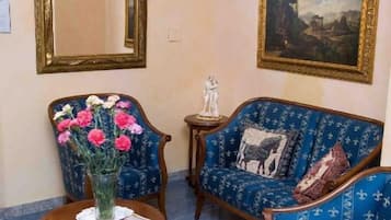 A room with a blue patterned sofa, a matching armchair, a small table with a vase of flowers, a framed painting, and a large mirror.