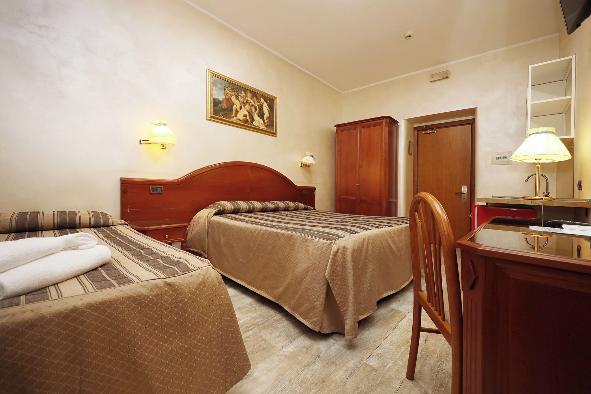 A hotel room with two beds, a wooden wardrobe, a desk with a lamp, and a painting on the wall.