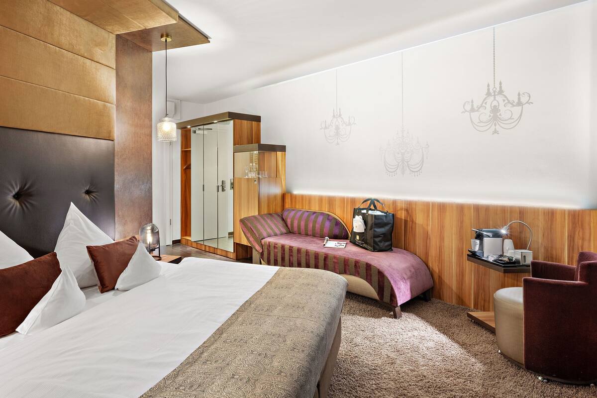 A modern hotel room with a large bed, a seating area, a wardrobe, and a desk with a lamp.