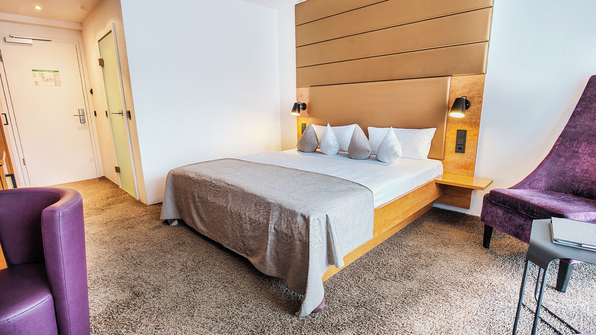 A hotel room with a bed, a purple armchair, a small table, and a wall-mounted light fixture.