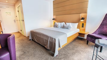 A hotel room with a bed, a purple armchair, a small table, and a wall-mounted light fixture.